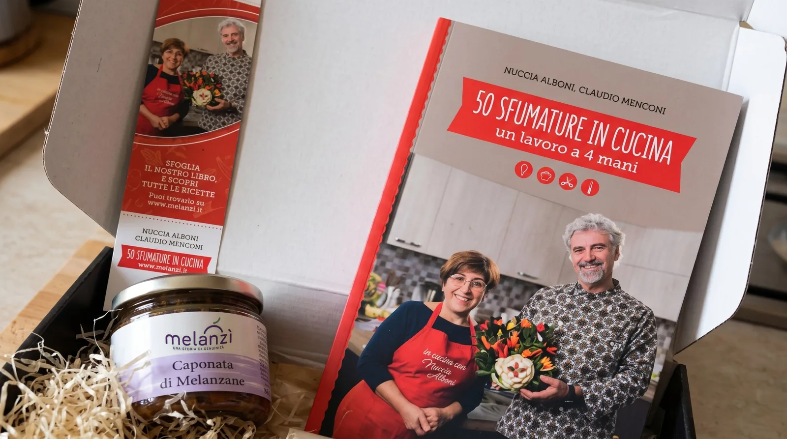 Box regalo aperta con il libro 50 sfumature in cucina, il segnalibro e un vasetto di caponata di melanzane Melanzì