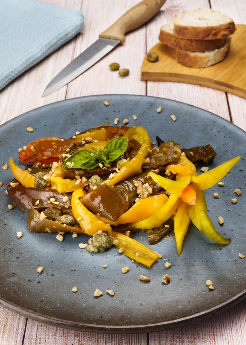 Peperoni ammollicati gialli e rossi con capperi e basilico su un piatto grigio, con fette di pane dietro